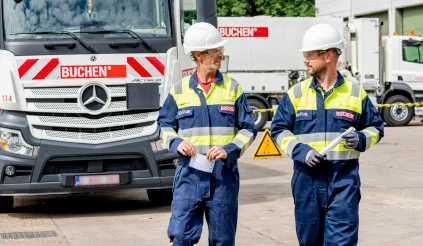 Twee collega’s van Buchen Industrial Services overleggen terwijl ze wandelen op een industriële locatie met vrachtwagens van Buchen op de achtergrond