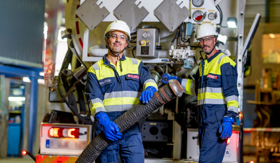 Twee operators industriële reiniging van Buchen Industrial Services staan achter een zuigwagen, waarbij de ene de zuigslang vasthoudt en de andere deze bedient.